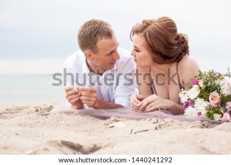 wedding couple sit in the beach and kissing against the sea or ocean. closeup of the bouquet lying next to the bride. woman wearing purple dress, the groom wearing white shirt