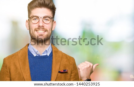 Young handsome business man wearing glasses smiling with happy face looking and pointing to the side with thumb up.