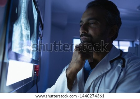 Side view of thoughtful mature mixed race male doctor looking at x-ray on x-ray light box in operation room at hospital