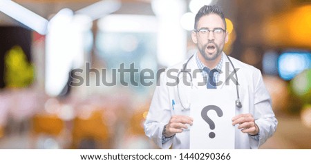 Handsome young doctor man holding paper with question mark over isolated background scared in shock with a surprise face, afraid and excited with fear expression