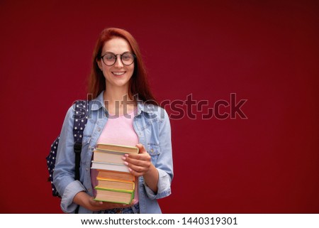 portrait of a beautiful girl student with a backpack and a stack of books in his hands is smiling at the red background. attractive positive the girl is a student of the senior classes in the glasses.