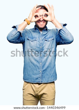 Handsome man wearing glasses doing ok gesture like binoculars sticking tongue out, eyes looking through fingers. Crazy expression.