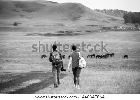 Two young girls with backpacks and photo camera on rural road. Horse farm pasture with mare and foal. Small village with old houses. Summer landscape with green hills.Black and white photo