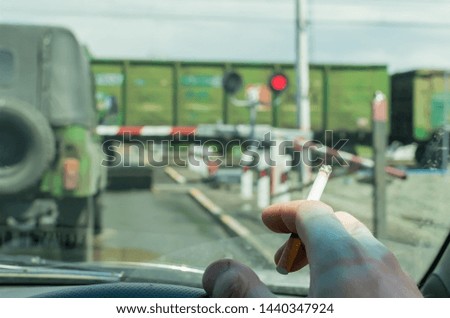 View of the driver hand with a cigarette on the steering wheel of the car, which stopped before a closed railway crossing at a red light