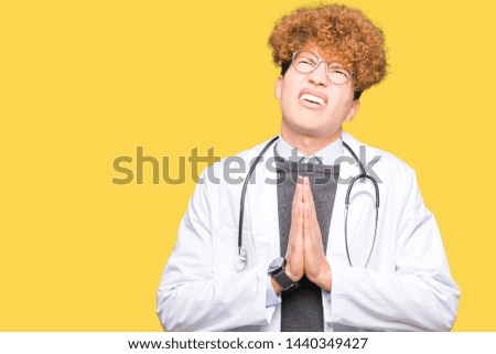 Young handsome doctor man wearing medical coat begging and praying with hands together with hope expression on face very emotional and worried. Asking for forgiveness. Religion concept.
