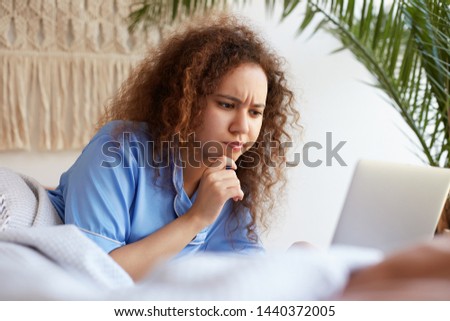 Close up of frowning young curly african american girl, dressed in blue pajamas and lying in bed, working at a laptop, doubtful looking at the monitor and touching cheek.