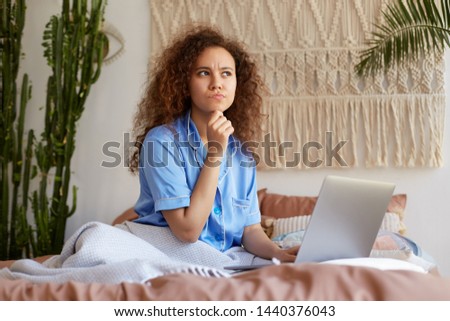 Photo of thinking young curly girl lying in bed, dressed in blue pajamas, working at a laptop, frowning, thoughtfully looking away and touching the chin.