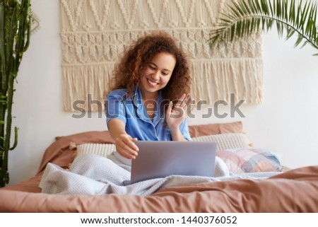Portrait of positive young curly girl siting in bed , dressed in blue pajamas, broadly smiling, looking at a laptop monitor and waving his hand, says hello to relatives through video chat.