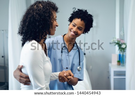 Front view of happy African American female doctor helping female patient to walk in the ward at hospital