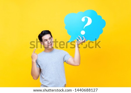 Attractive Asian man holding question mark bubble sign isolated on yellow studio background