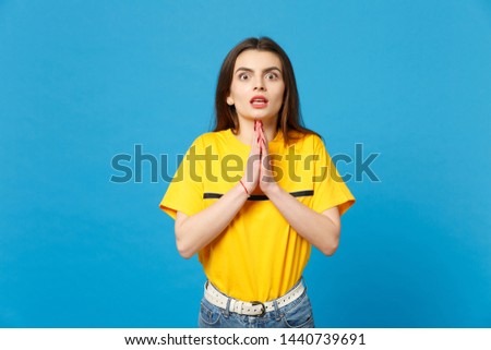Portrait of shocked young woman in vivid casual clothes looking camera, holding hands folded, praying isolated on bright blue wall background in studio. People lifestyle concept. Mock up copy space