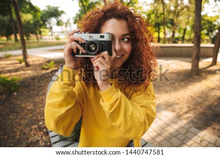 Happy young girl with red curly hair sitting on a bench at the park, taking pictures with photo camera