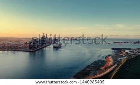 Sunrise over Felixstowe Container Port as two tugs shepherd a container ship from under blue gantry cranes with rows of shipping containers stacked behind them to the right Harwich and Shotley Marina Royalty-Free Stock Photo #1440965405