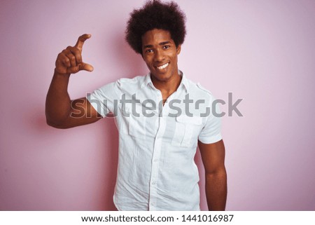 Young american man with afro hair wearing white shirt standing over isolated pink background smiling and confident gesturing with hand doing small size sign with fingers looking and the camera