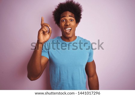 African american man with afro hair wearing blue t-shirt standing over isolated pink background pointing finger up with successful idea. Exited and happy. Number one.