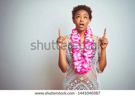 Young african american woman with afro hair wearing flower hawaiian lei over isolated background amazed and surprised looking up and pointing with fingers and raised arms.