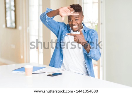 African american student man studying using notebooks and wearing headphones smiling making frame with hands and fingers with happy face. Creativity and photography concept.