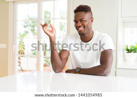 Handsome african american man on white table at home with a big smile on face, pointing with hand and finger to the side looking at the camera.