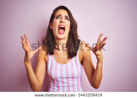 Young beautiful woman wearing striped pink swimsuit swimwear over pink isolated background crazy and mad shouting and yelling with aggressive expression and arms raised. Frustration concept.