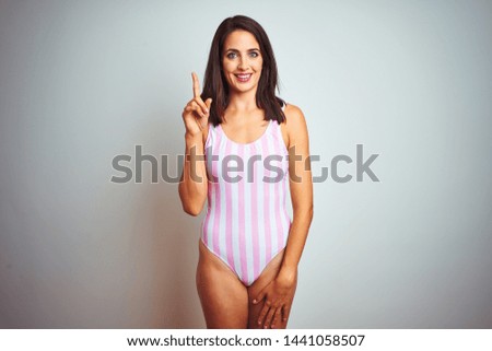 Young beautiful woman wearing striped pink swimsuit swimwear over isolated background showing and pointing up with finger number one while smiling confident and happy.