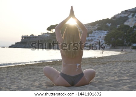 Backlighting image of a blonde girl sits in a yoga position back to the camera and raising arms