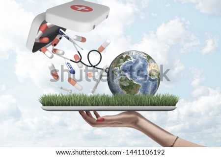 Female hand holding earth globe with pills, syringes and digital thermometers falling out of first aid medical kit on green grass model with blue sky background. Ecology and environment. Environmental