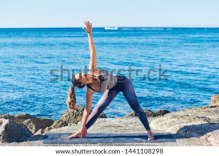 Beautiful young woman practicing yoga on seaside against blue sea. Athletic lady doing triangle pose on the beach. Healthy lifestyle.