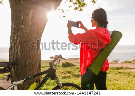 slim fit beautiful woman doing sports in morning in park riding on bicycle with yoga mat in colorful fitness outfit, making photo on phone, exploring natures, miling happy healthy lifestyle