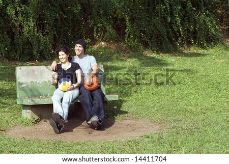 A couple is sitting on a park bench.  They are both smiling and laughing and looking away from the camera and each other. Horizontally framed photo.