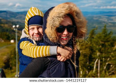 Happy woman holds her son on her back on a background of mountains in autumn