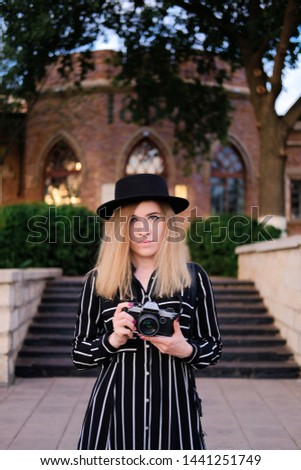 Blonde girl in black hat taking pictures with retro design camera on background of historic building. City traveling alone concept.
