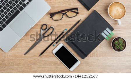 
On the office desk, a smartphone with a blank screen, a laptop, glasses, notepads, scissors, pencils, a cactus and a cup of coffee. Top view.Mockup.
