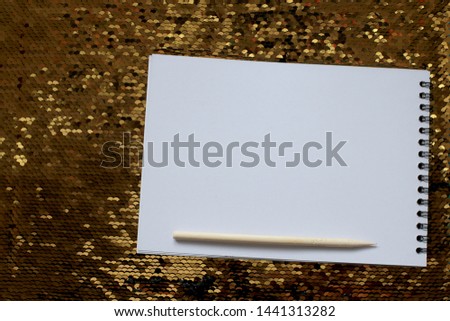 A notebook with empty white sheets and a wooden scraper are on a bright shiny background of gold sequins, a place to write notes, write a reminder text on white paper