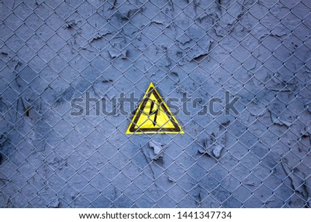 old gray blue wall with peeling paint under a metal grid with a triangular yellow sign of dangerous electrical voltage. rough surface texture