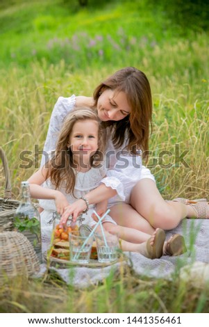 Beautiful young mother and her little daughter in white dress having fun in a picnic. They sit on the rug and take the berries out of the jar. Maternal care and love. Verticalal photo.