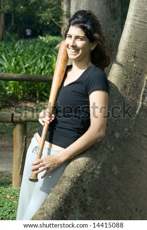 A young, attractive woman is leaning against a tree at a park.  She is smiling and looking at the camera.  She is posing with a baseball bat.  Vertically framed photo.