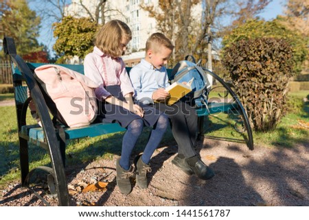 Little boy and girl schoolchildren reading book, sitting on bench, children with backpacks, bright sunny autumn day