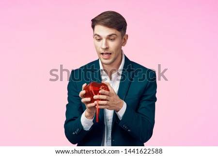  A man in a business suit on a pink background holds in his hand a gift box in the shape of a heart                  