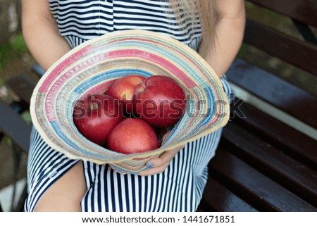 A girl sits on a bench and holds red apples with a hat