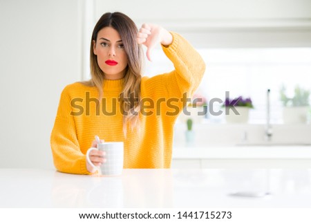 Young beautiful woman drinking a cup of coffee at home with angry face, negative sign showing dislike with thumbs down, rejection concept
