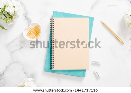 Modern home office desk workspace with blank paper notebook, white flowers and feminine accessories on marble background. Flat lay, top view, overhead. Woman workspace concept. 