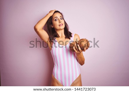 Beautiful woman wearing pink striped swimsuit holding coconuts over isolated pink background stressed with hand on head, shocked with shame and surprise face, angry and frustrated. Fear and upset