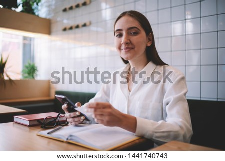 Positive smiling hipster girl sitting at coffee shop while looking at camera and holding business card in hand, portrait of woman using visit card for contact via mobile phone  spending time at cafe