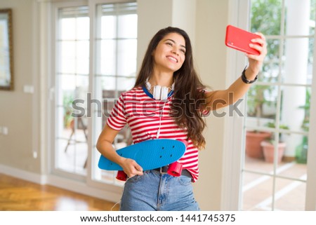 Beautiful skater woman smiling friendly standing with skateboard and taking a selfie using smartphone