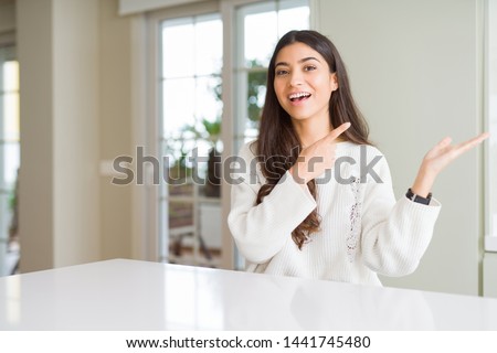 Young beautiful woman at home on white table amazed and smiling to the camera while presenting with hand and pointing with finger.