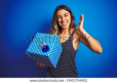 Young woman holding birthday present over blue isolated background very happy and excited, winner expression celebrating victory screaming with big smile and raised hands