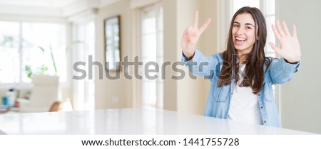 Wide angle picture of beautiful young woman sitting on white table at home showing and pointing up with fingers number seven while smiling confident and happy.