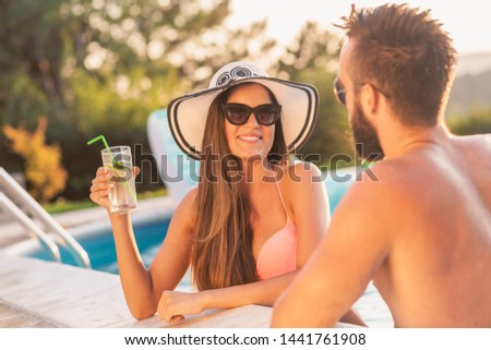 Couple in love at a poolside summer party, standing in water next to the swimming pool edge, drinking beer and having fun
