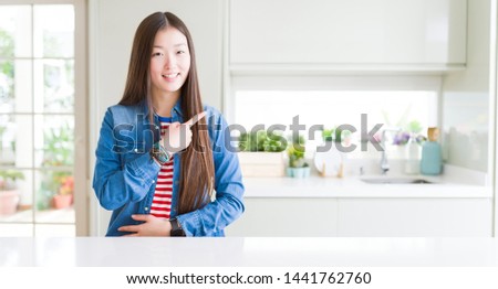 Beautiful Asian woman wearing denim jacket on white table cheerful with a smile of face pointing with hand and finger up to the side with happy and natural expression on face