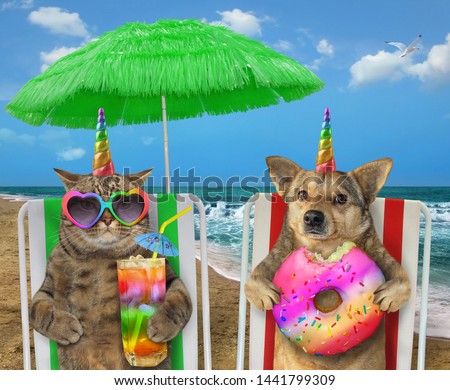 The dog unicorn with a donut and the cat in sunglasses with a glass of colored cocktail are sitting under the green straw umbrella on a beach chairs on the sea shore.